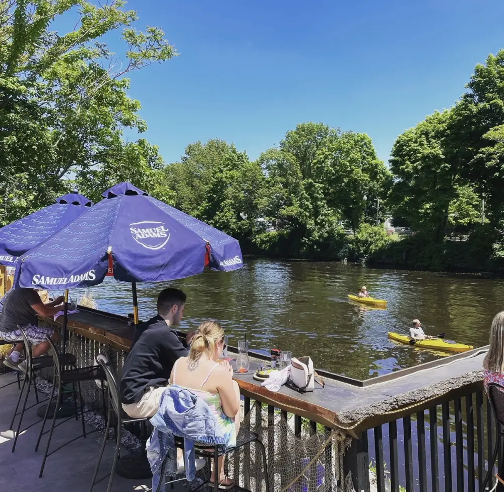 Diners at the Riverhouse riverside deck with kayakers paddling past on the Taunton River